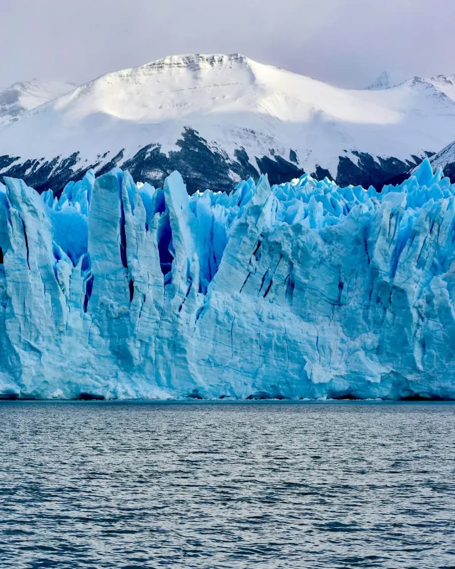 Blau schimmernde Gletscherfront vor einem schneebedeckten Berg am Wasser